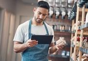 A man in an apron stands next to shelves of products with a tablet in one hand and a jar in the other.