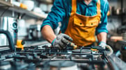 A worker wearing a protective apron and heavy gloves works on a gas range.
