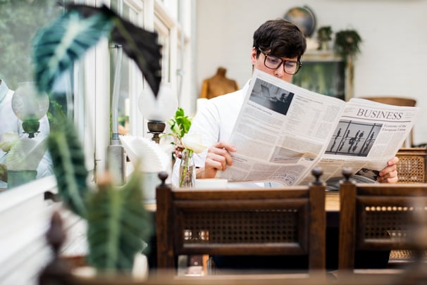 A man wearing glasses reads the business section of a newspaper sitting in a restaurant. 