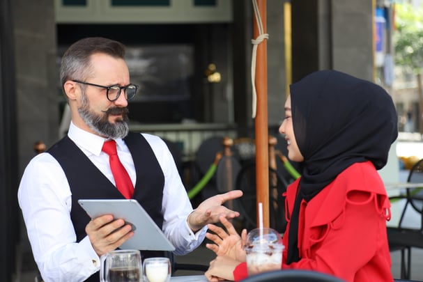 A man dressed like a waiter in a black vest and red tie holds a tablet as he talks to a woman in a hijab.  