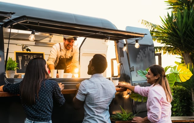 A smiling man in an apron talks to three customers outside his food truck.  