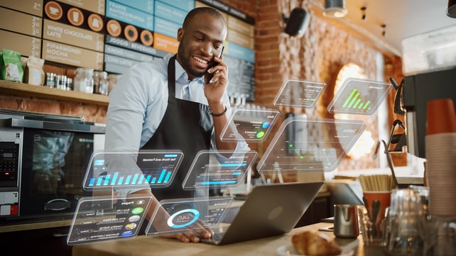 A man stands behind a cafe counter using a laptop; images indicate that he is looking at an array of data. 