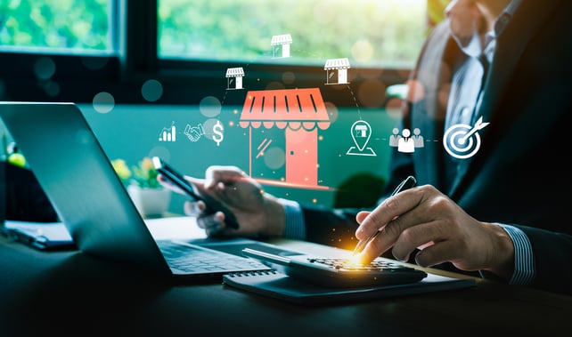 A person is sitting at a laptop with a phone in one hand and a calculator in the other; floating icons indicate they are looking at marketing data. 