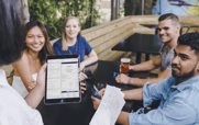 A server holding a tablet with an order on it stands next to a table of smiling diners.